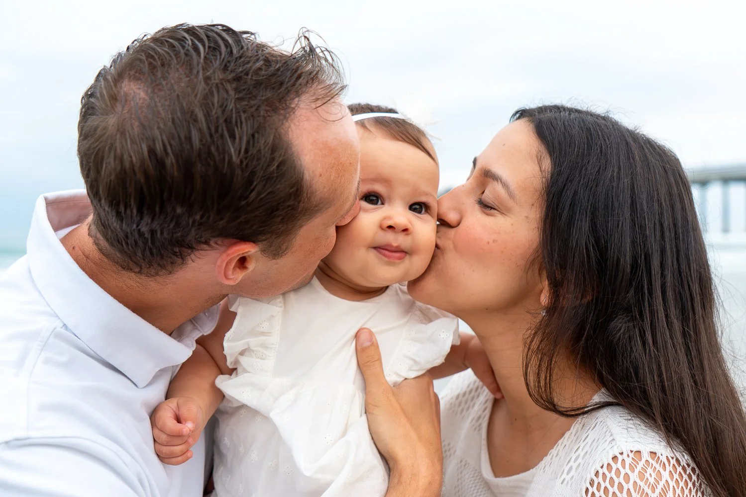A family of three, consisting of a father, mother, and baby, sharing a moment of affection outdoors with blurred background.