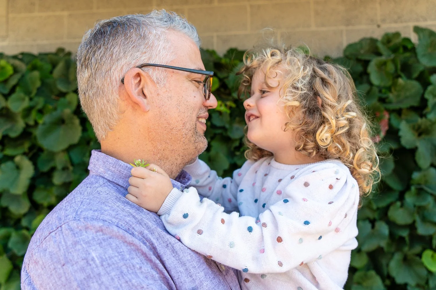 A middle-aged man with glasses and a purple shirt holding a young girl with curly blonde hair and a white polka dot shirt, both smiling and looking at each other outdoors with green foliage in the background.