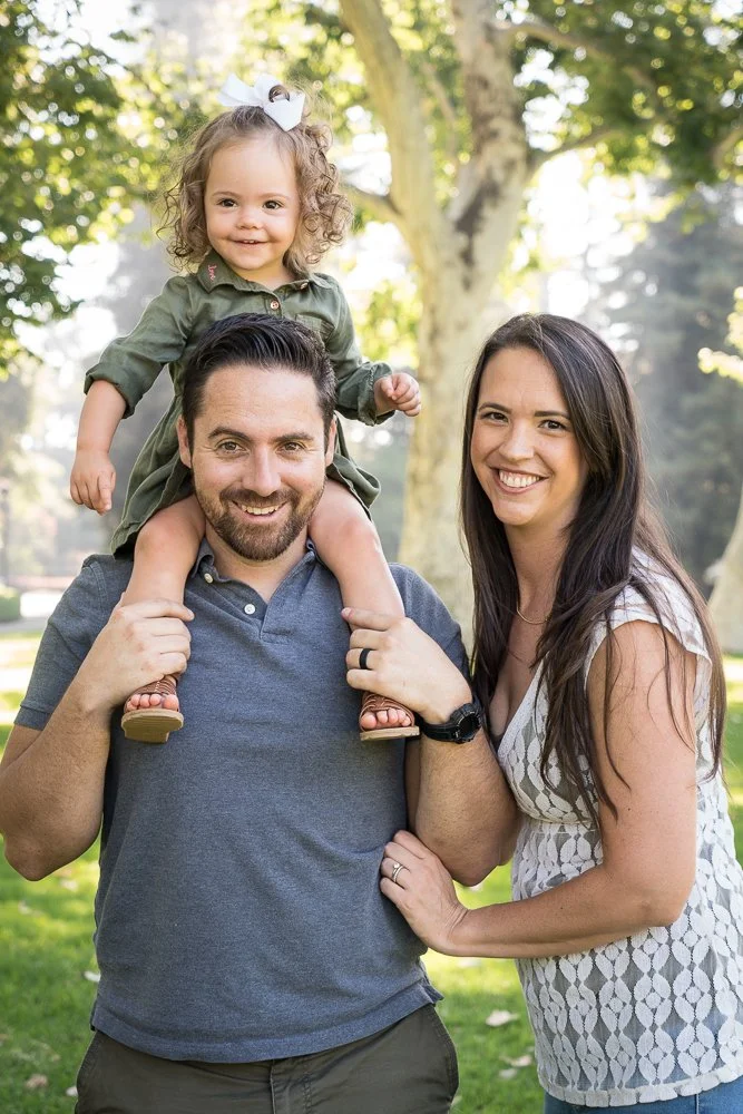 A family of three outdoors in a park with trees, featuring a smiling man carrying a young girl on his shoulders and a smiling woman standing beside them.