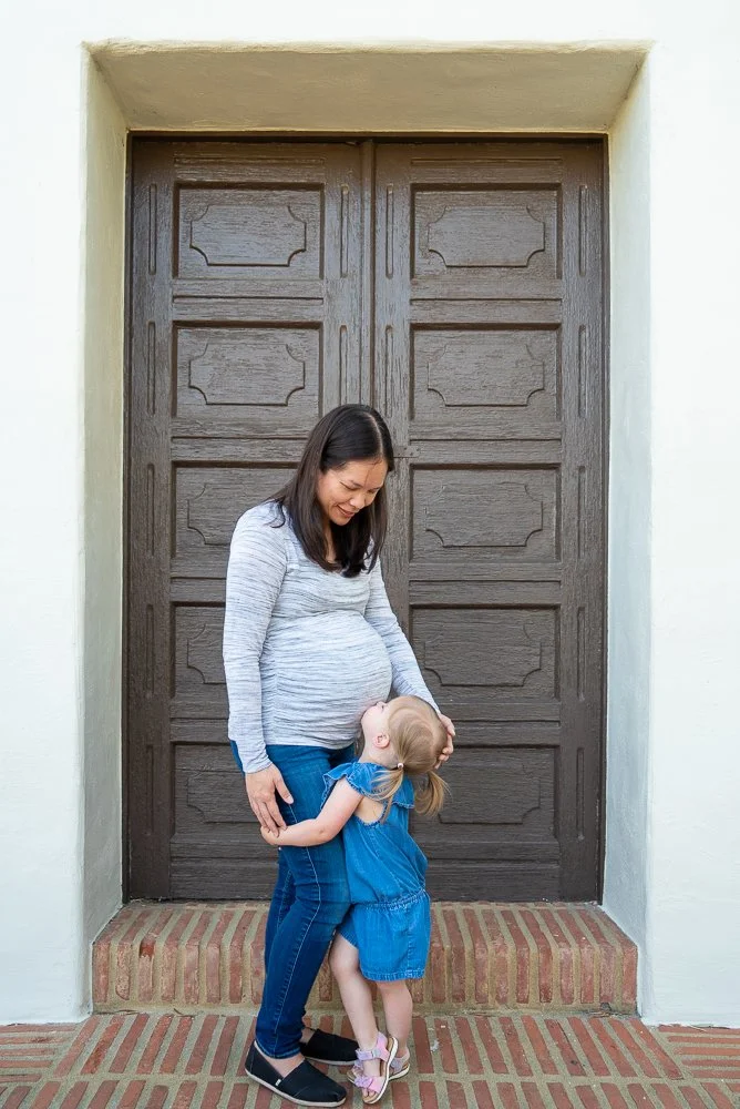 A pregnant woman and a young girl hugging in front of a large wooden door with brick steps.