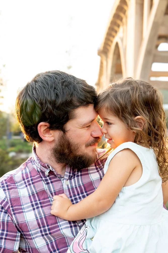 A father and daughter sharing a joyful moment with their foreheads touching outdoors near a bridge during sunset.