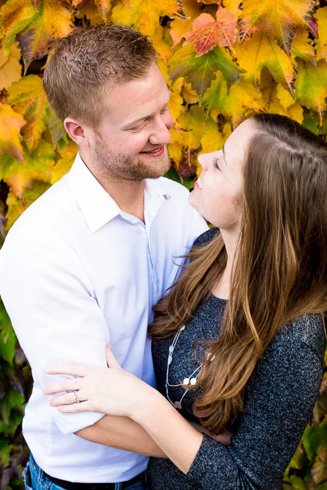 A man and a woman embracing outdoors with colorful autumn leaves in the background.