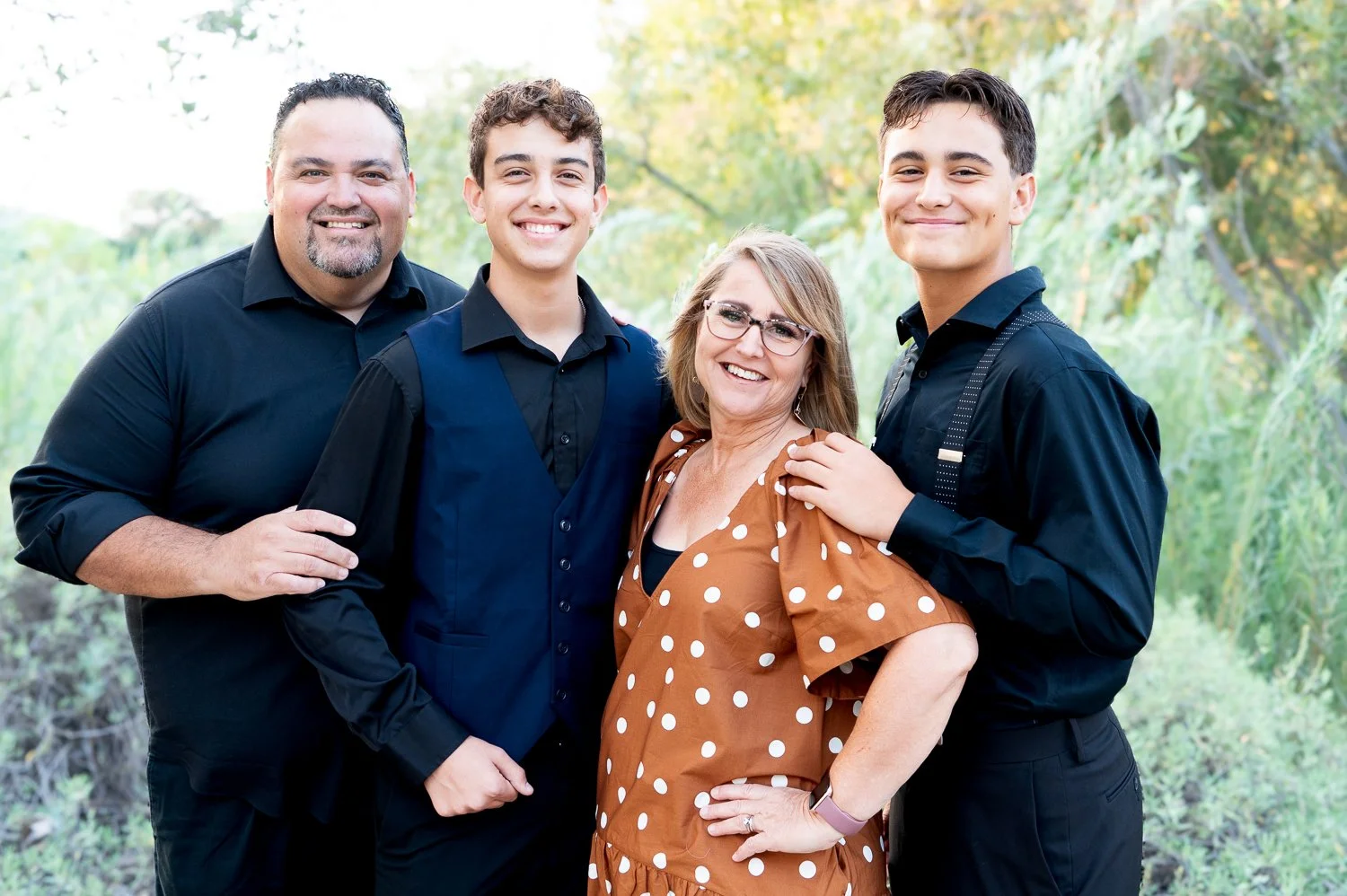 Family portrait of four people outdoors, two adults and two teenagers, smiling and standing close together with greenery in the background.