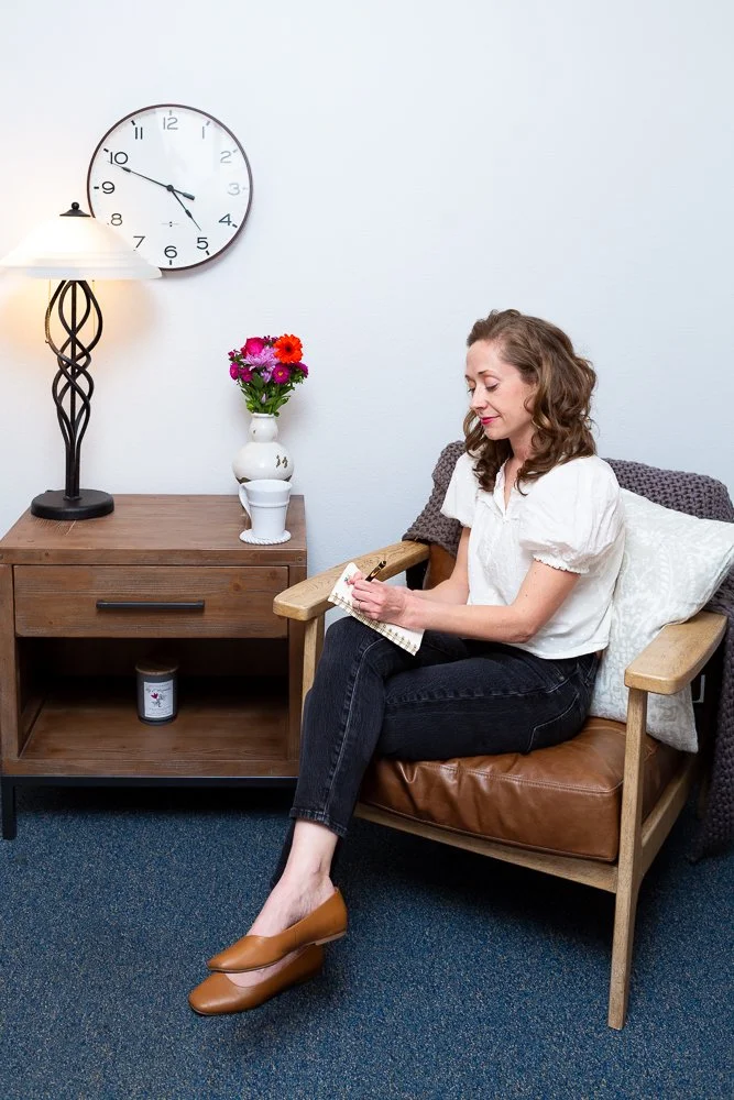 A woman sitting on a wooden armchair with brown leather cushion, writing in a notebook. She is wearing a white blouse, black jeans, and brown shoes. Next to her is a side table with a white vase holding pink and purple flowers, a white mug, and a can