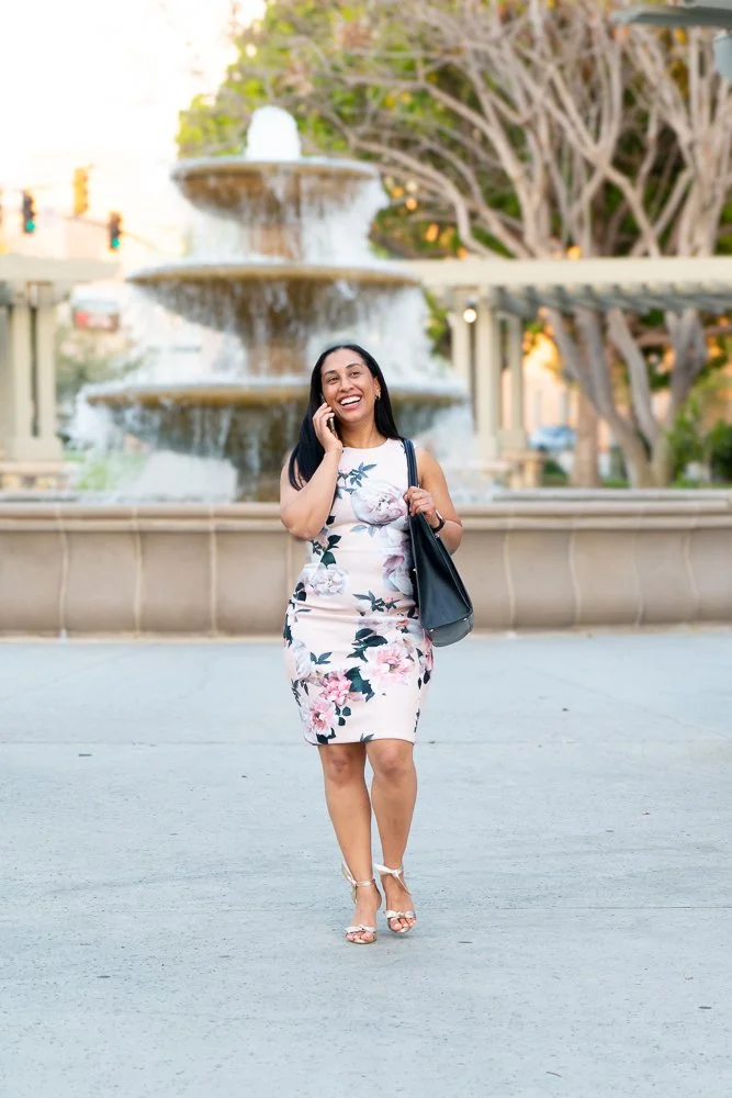 A woman in a floral dress smiling and talking on her phone while walking in front of a fountain in an outdoor urban setting.
