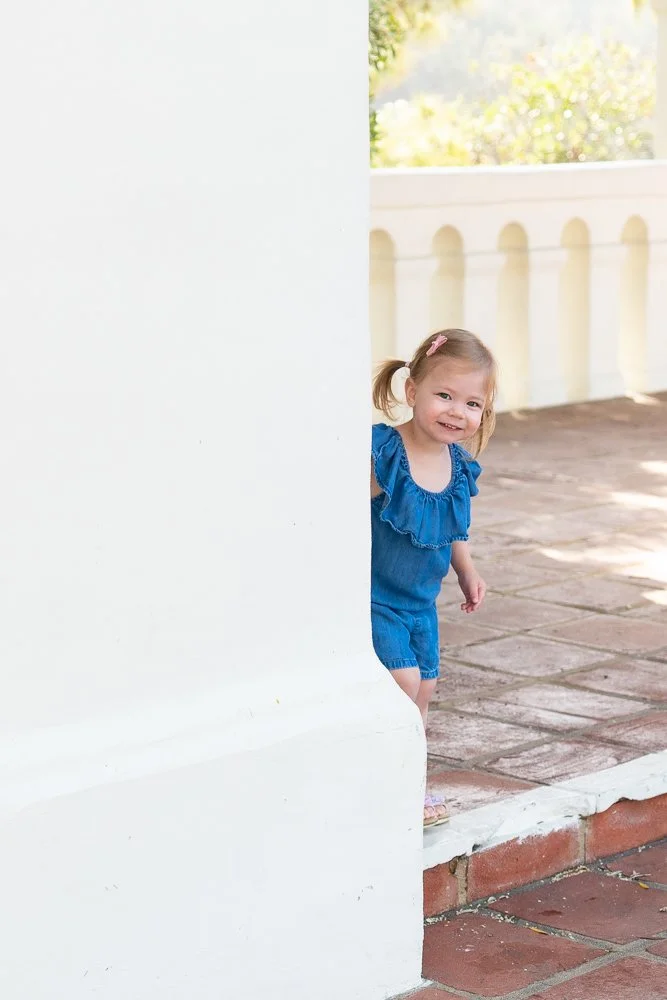 A young girl with pigtails and a pink hair clip, wearing a blue dress, peeking out from behind a white wall and smiling.