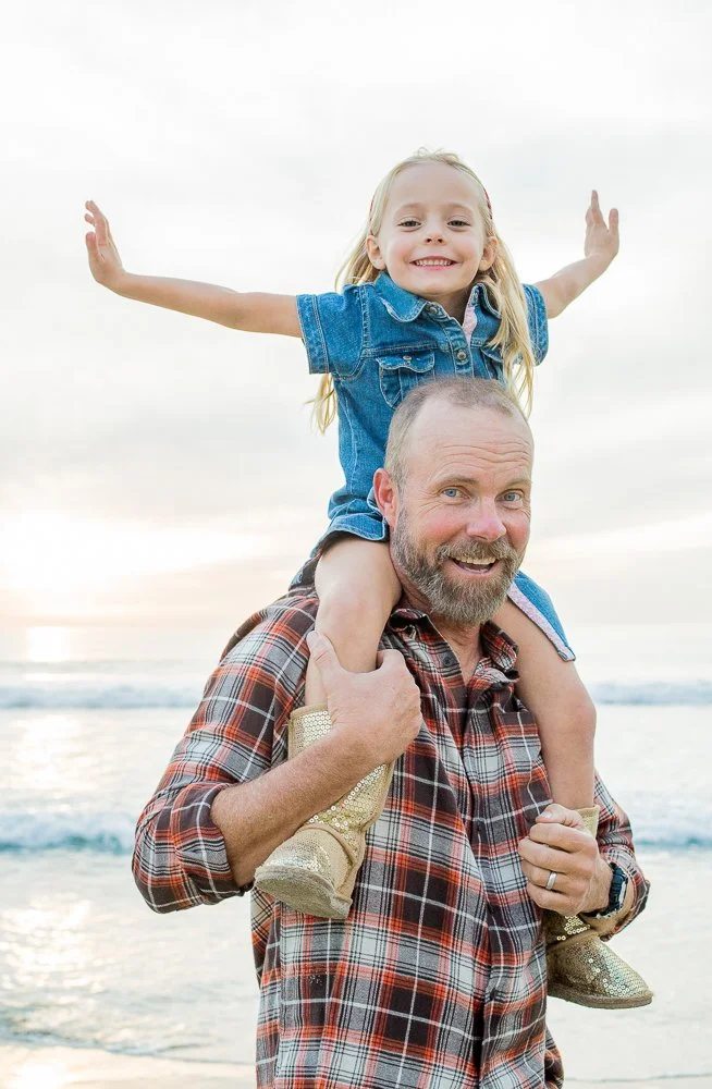 A man carries a young girl on his shoulders on a beach during sunset, both smiling.