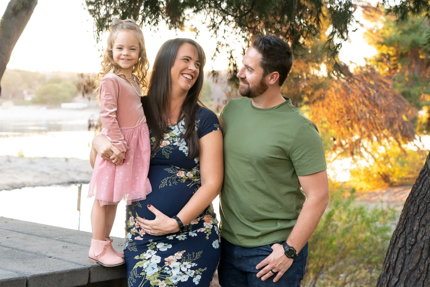 A family of three enjoying outdoor fall scenery, a woman holding a young girl on her lap, the woman is pregnant, all smiling at each other, with trees and a body of water in the background.