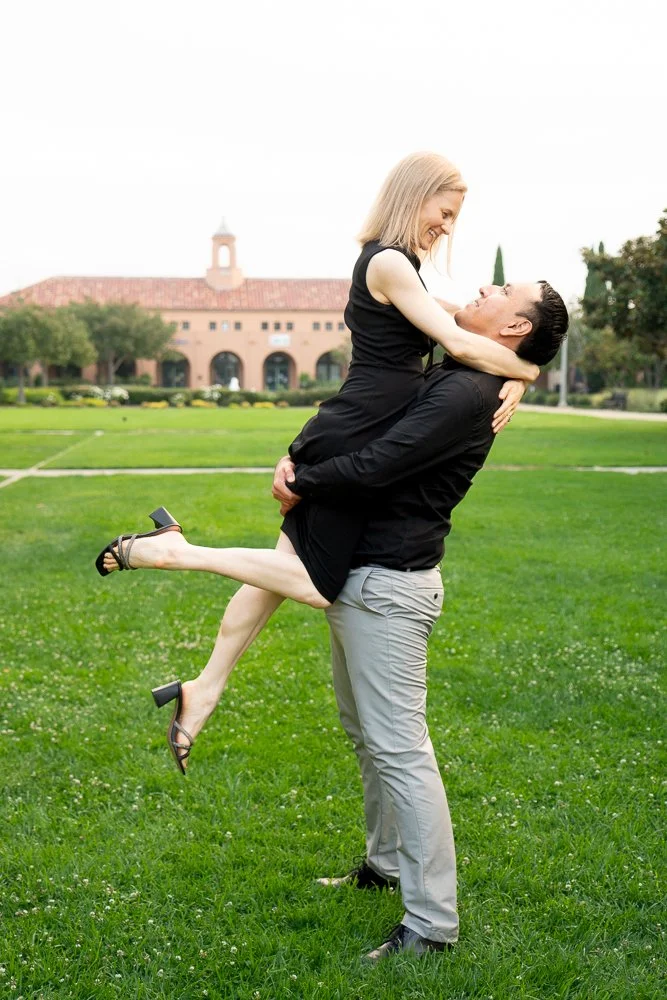 A man lifting a woman in a park with a building in the background