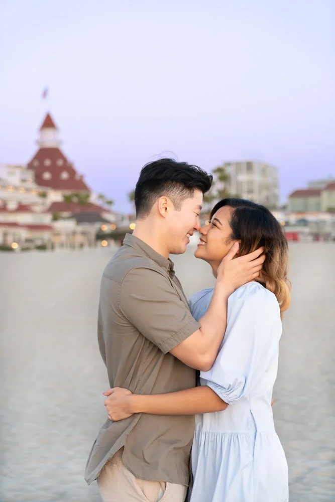 A couple embracing on the beach, faces close together smiling, with a blurred background of buildings and a tower.