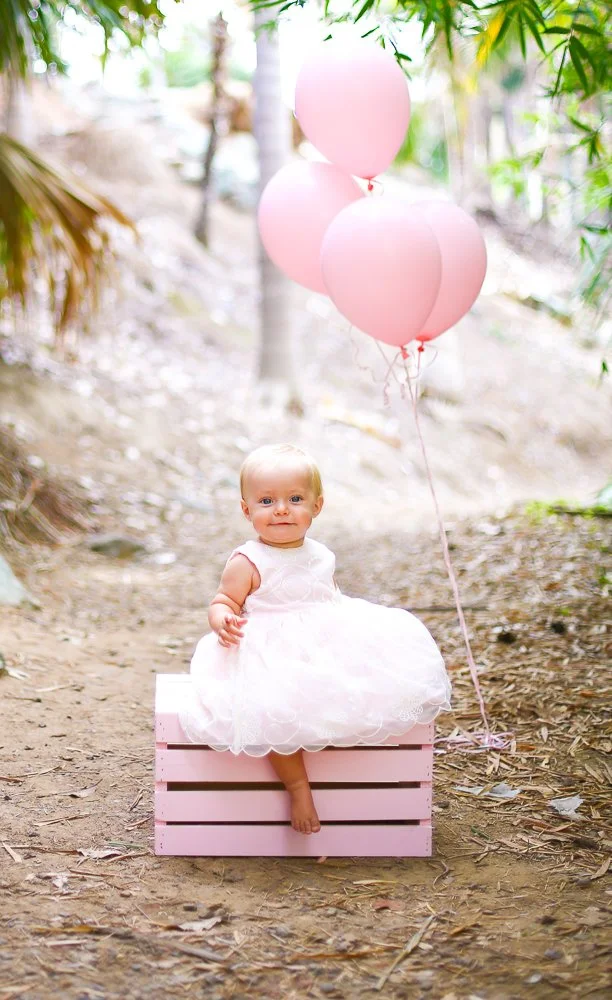 A young girl in a white dress sitting on a pink wooden crate outdoors, holding three pink balloons on strings, surrounded by trees and greenery.