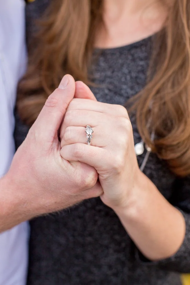Close-up of a woman’s hand with a diamond engagement ring, held by another person, in front of her chest with brown hair and casual clothing.