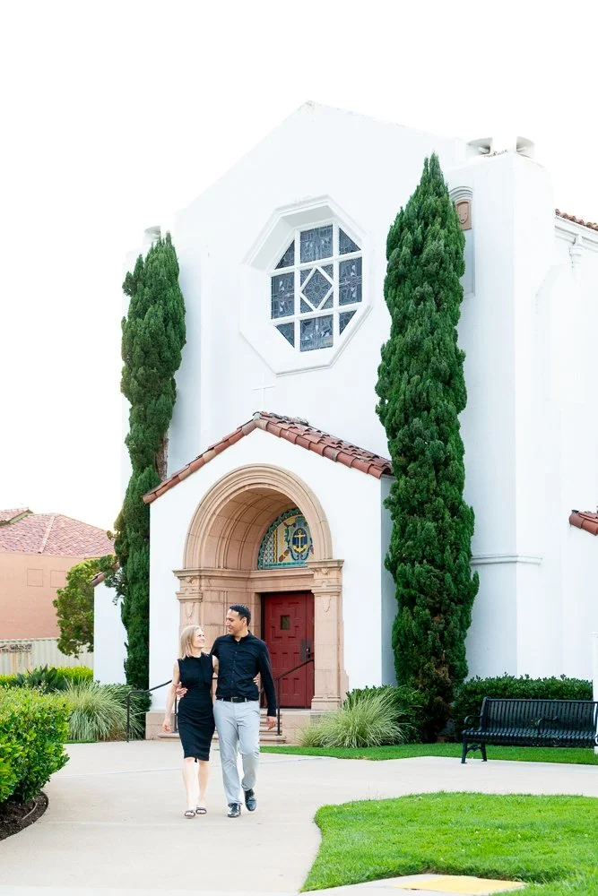 Two people walking on sidewalk near a white church with tall green trees, a red door, and a decorative window above the entrance.