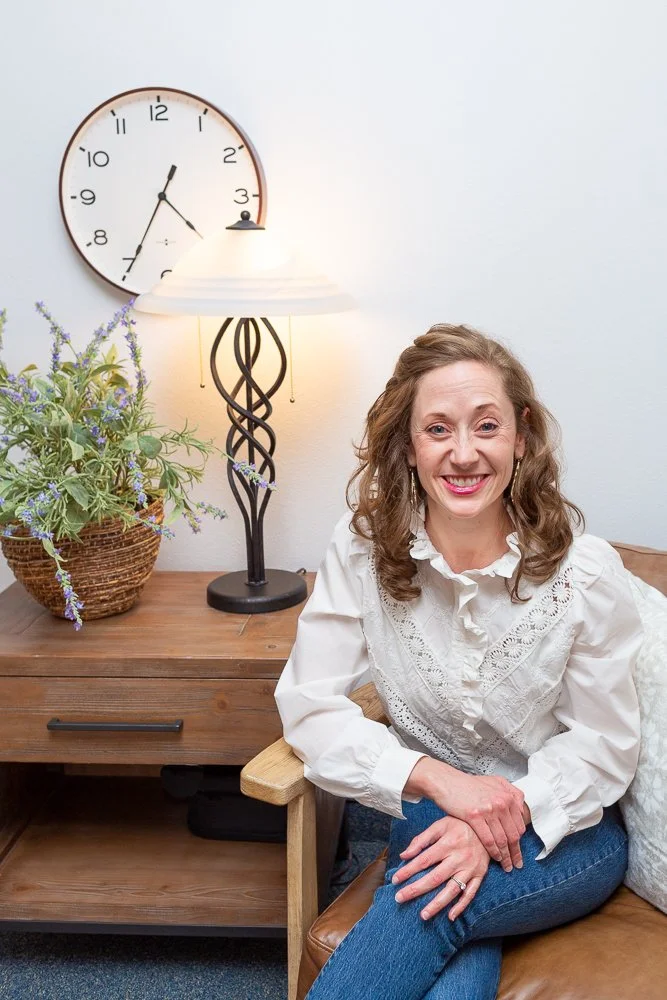 Smiling woman sitting on a brown leather chair in a cozy room, with a wooden side table, a decorative lamp, a large wall clock, and a potted plant with purple flowers in the background.