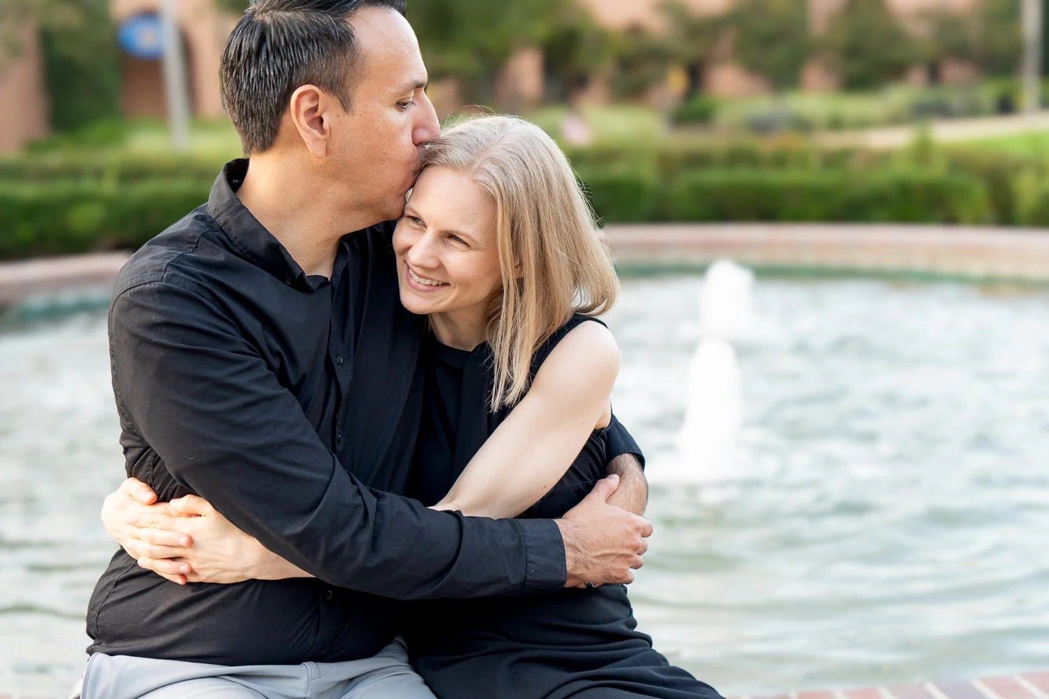 A man and woman hugging near a fountain in a park, with the man kissing the woman's head and the woman smiling, both wearing black clothing.