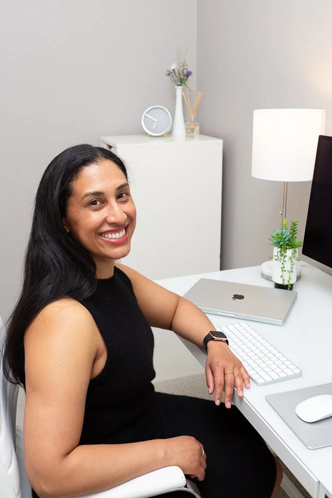 A woman with long dark hair smiling and sitting at a white desk with a computer, a closed MacBook, a keyboard, and a mouse. There is a lamp, a small potted plant, and a decorative vase with flowers in the background.