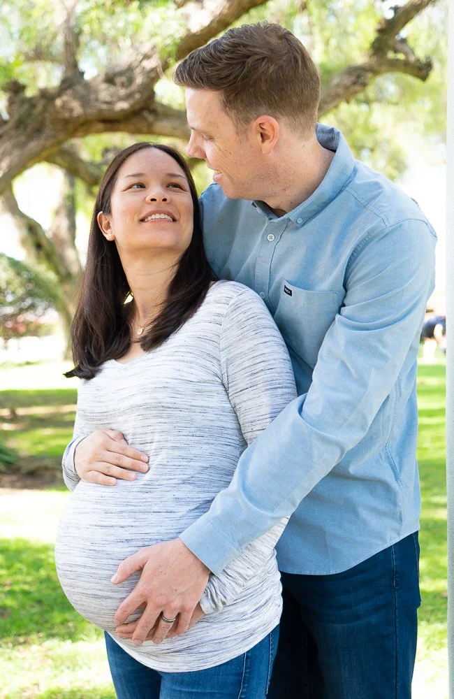 A pregnant woman with long dark hair smiling happily and looking at a man, while the man gazes down at her affectionately outdoors under a tree.