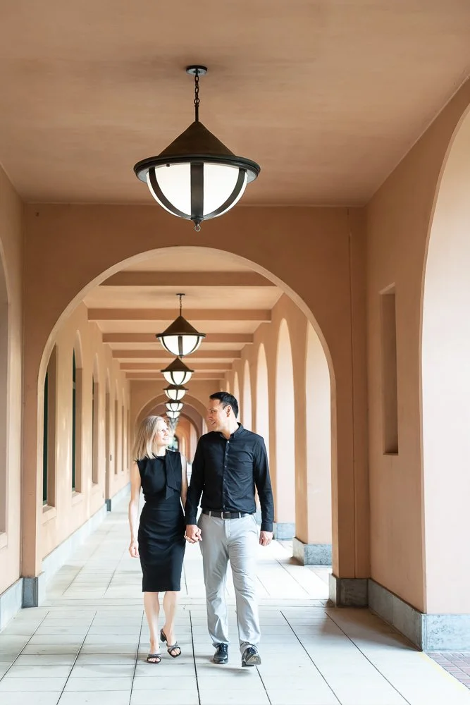 A couple walking hand in hand through an arched corridor with hanging lights and beige walls.