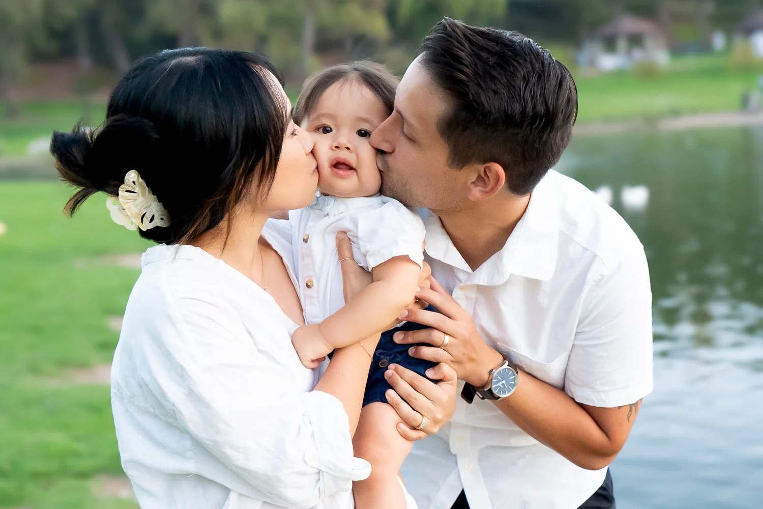 Family of three outside near a lake, with parents kissing their baby on the cheeks.