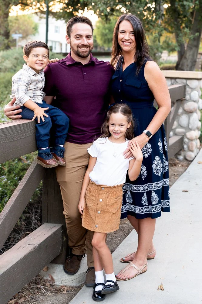 A family of four posing outdoors near a wooden fence with trees and houses in the background, during daytime. The father, mother, son, and daughter are smiling at the camera.