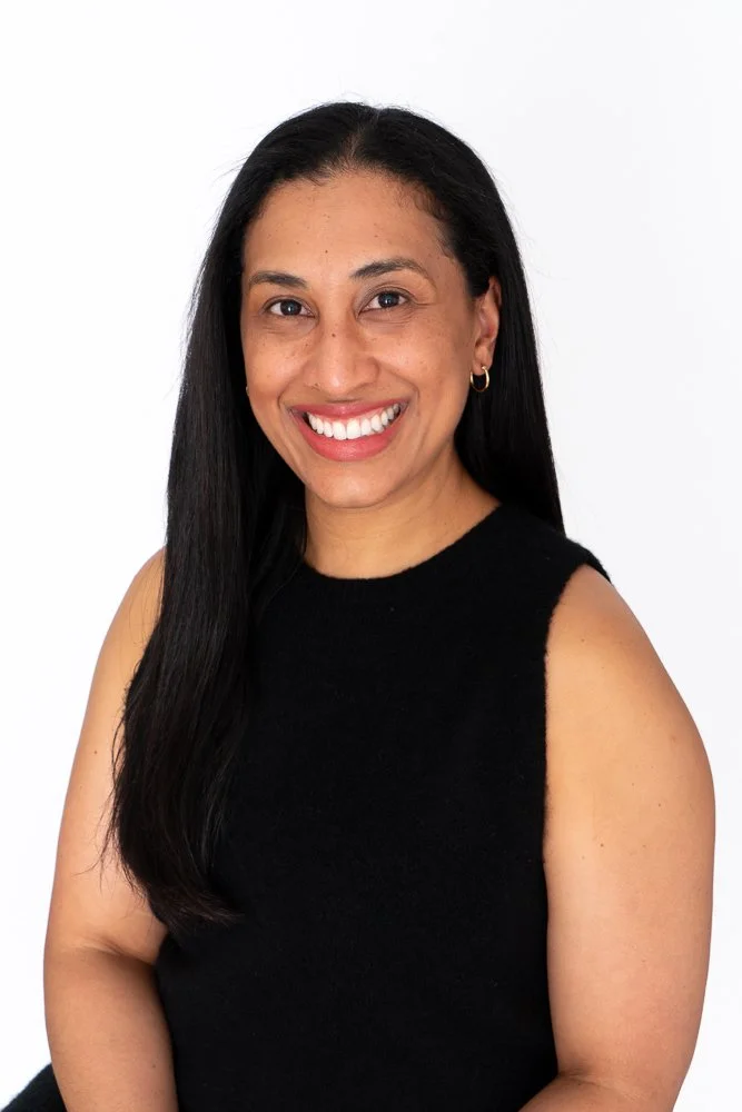 A smiling woman with long black hair wearing a sleeveless black top, earrings, and minimal makeup, posing against a white background.