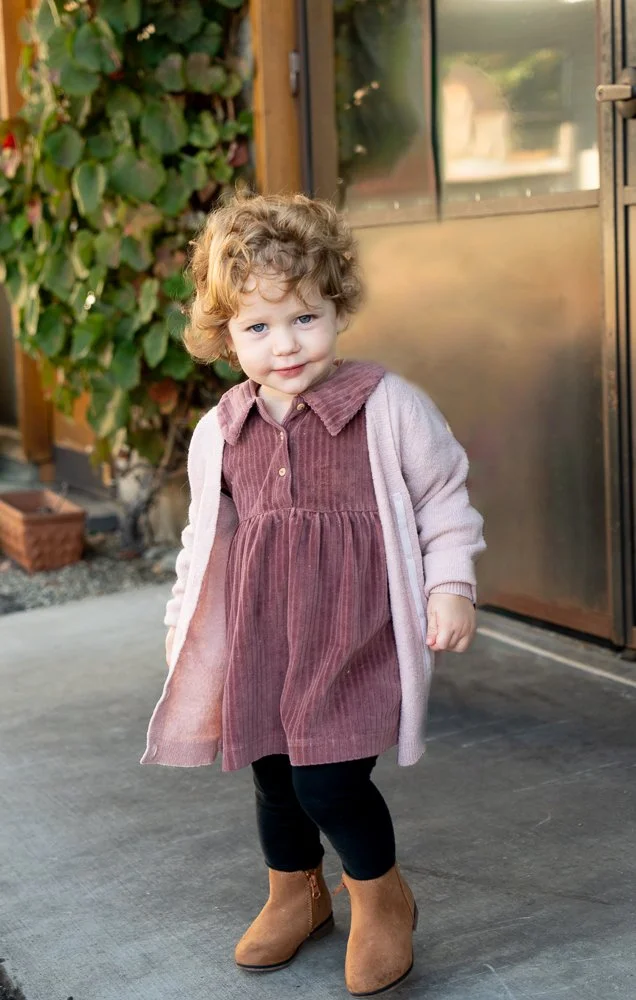 A young girl with curly blonde hair standing outside, wearing a pink dress, a light pink cardigan, black leggings, and brown ankle boots.