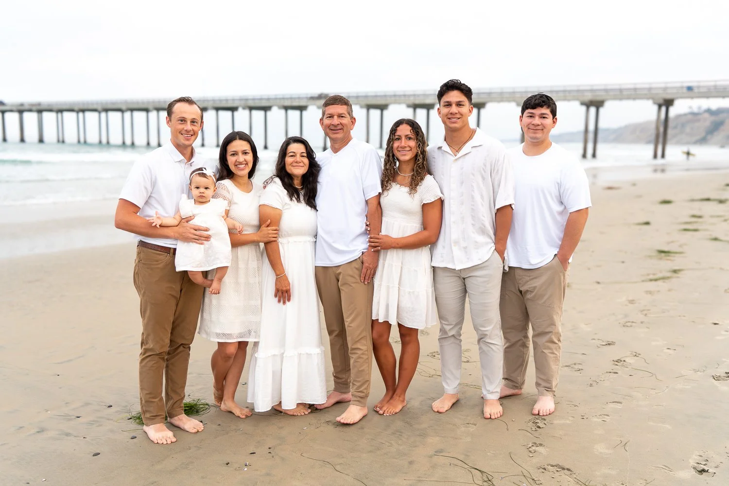 A large family, including children, standing on a sandy beach with an ocean and a pier in the background, all dressed in white or light-colored clothing.