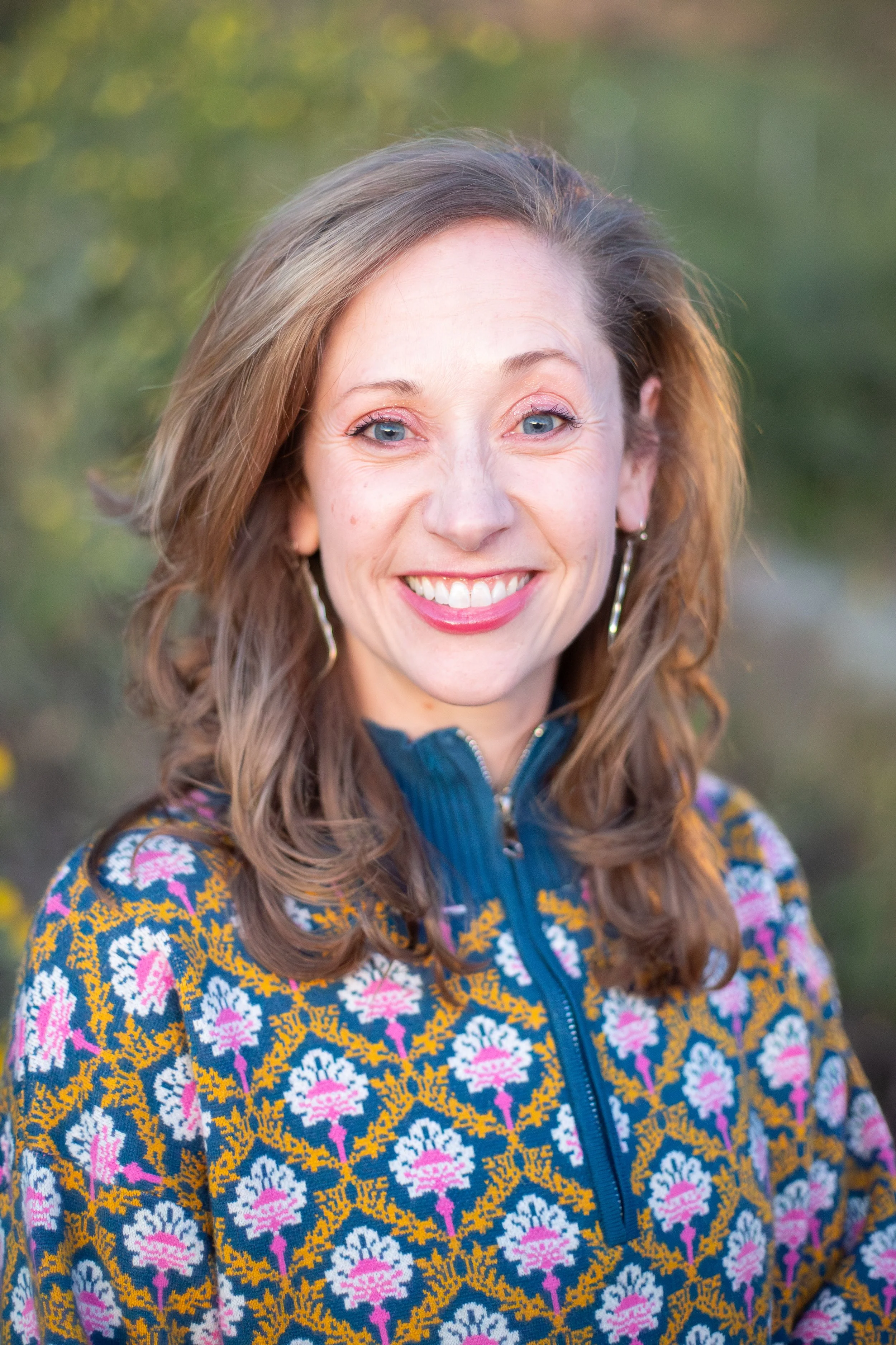 A woman with light brown wavy hair, blue eyes, and a wide smile wearing a patterned jacket in a natural outdoor setting.