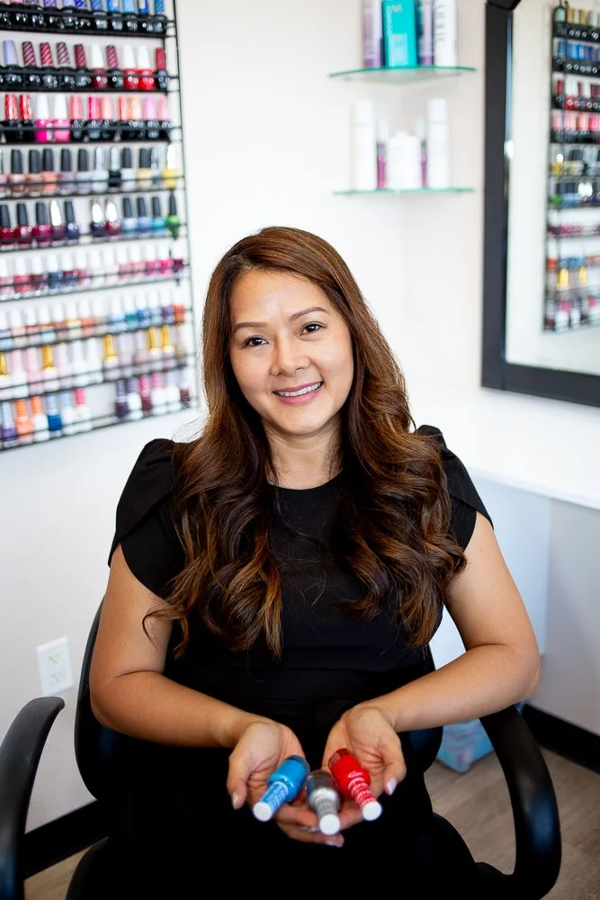 A woman with long brown hair sitting in a nail polish salon, holding two bottles of nail polish, one red and one blue.