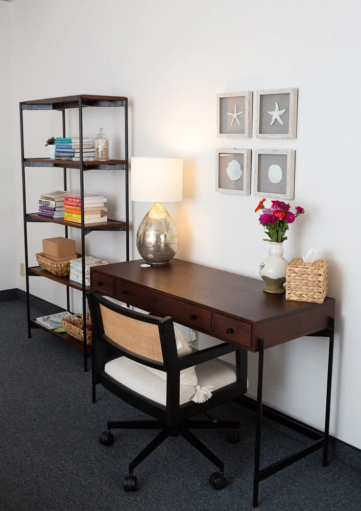 Home office desk with a chair, a table lamp, a flower vase with pink and purple flowers, a tissue box, and a bookshelf with books and baskets, against a white wall with framed starfish and shell artwork.