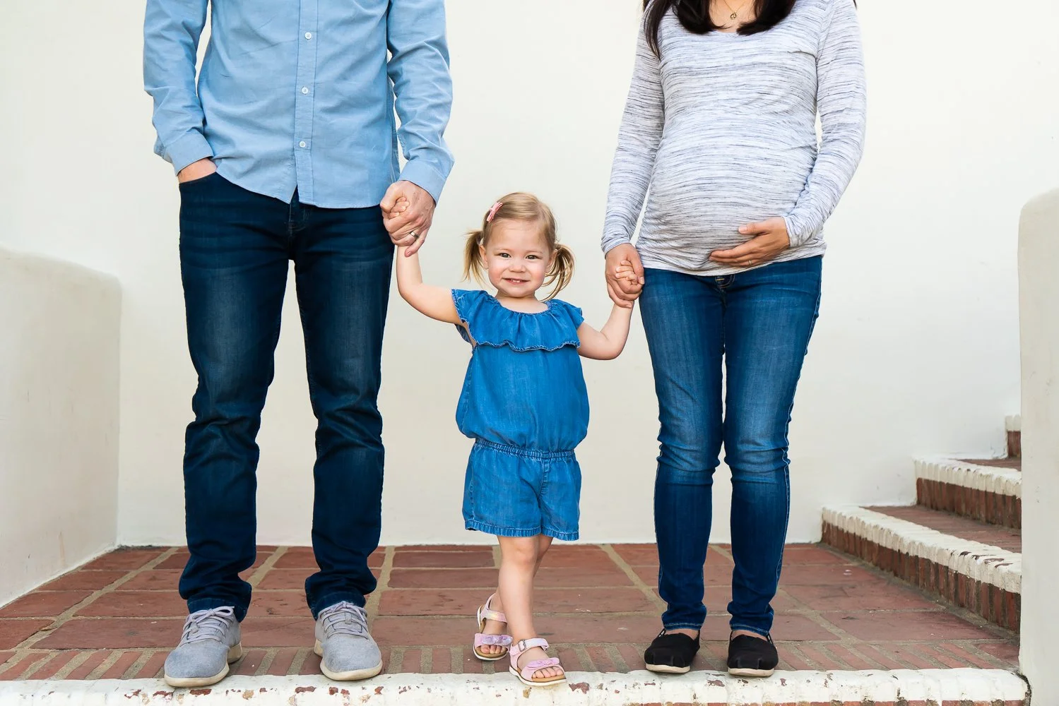 A young girl in a blue dress holding hands with a man and a pregnant woman, standing on brick steps against a plain wall.