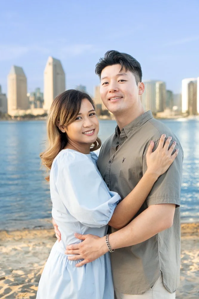 A smiling couple hugging on a beach with a city skyline and water in the background.