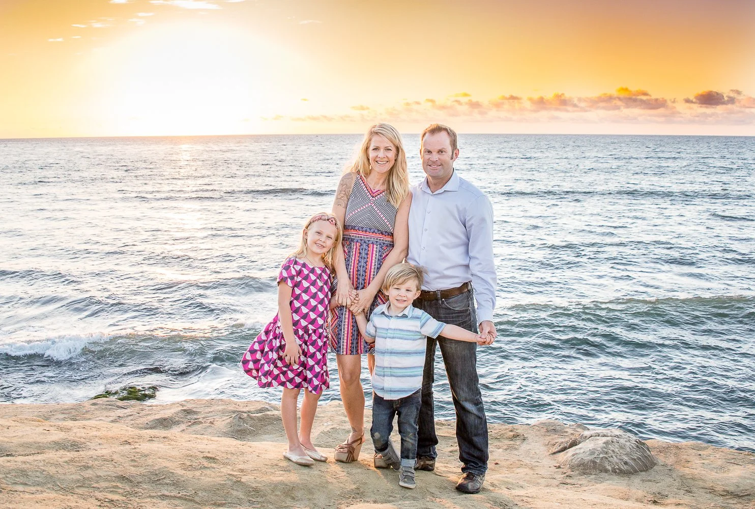 A family of four standing on a rocky beach at sunset, smiling at the camera. The background shows the ocean and a vibrant sky with pink, orange, and yellow hues.