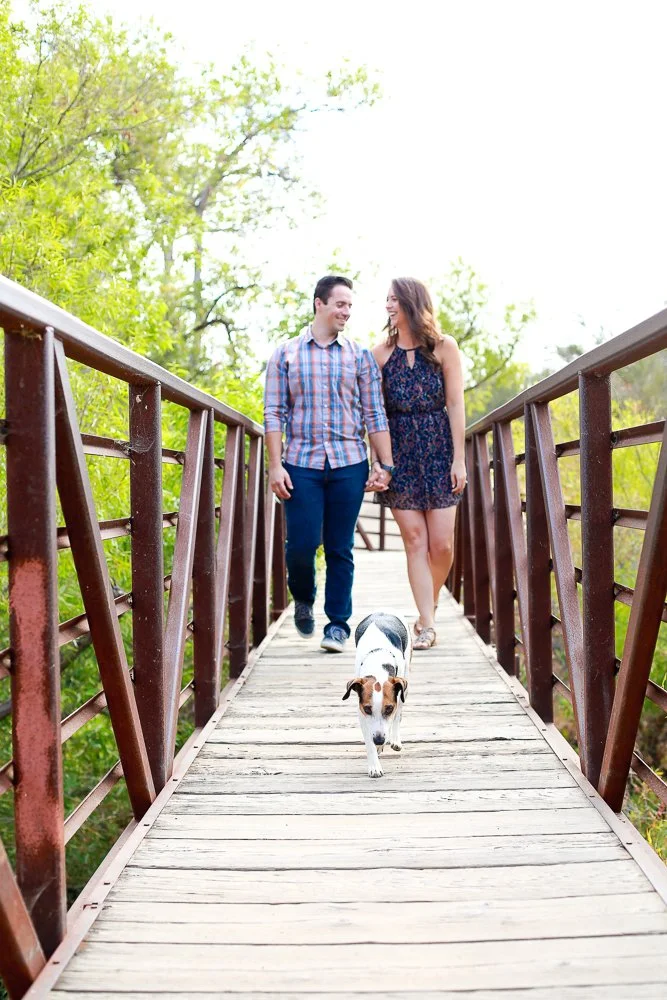 A couple walks hand in hand with their dog on a wooden bridge surrounded by green trees on a bright day.
