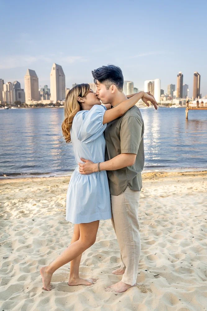 A couple kissing on a sandy beach with a city skyline and water in the background.