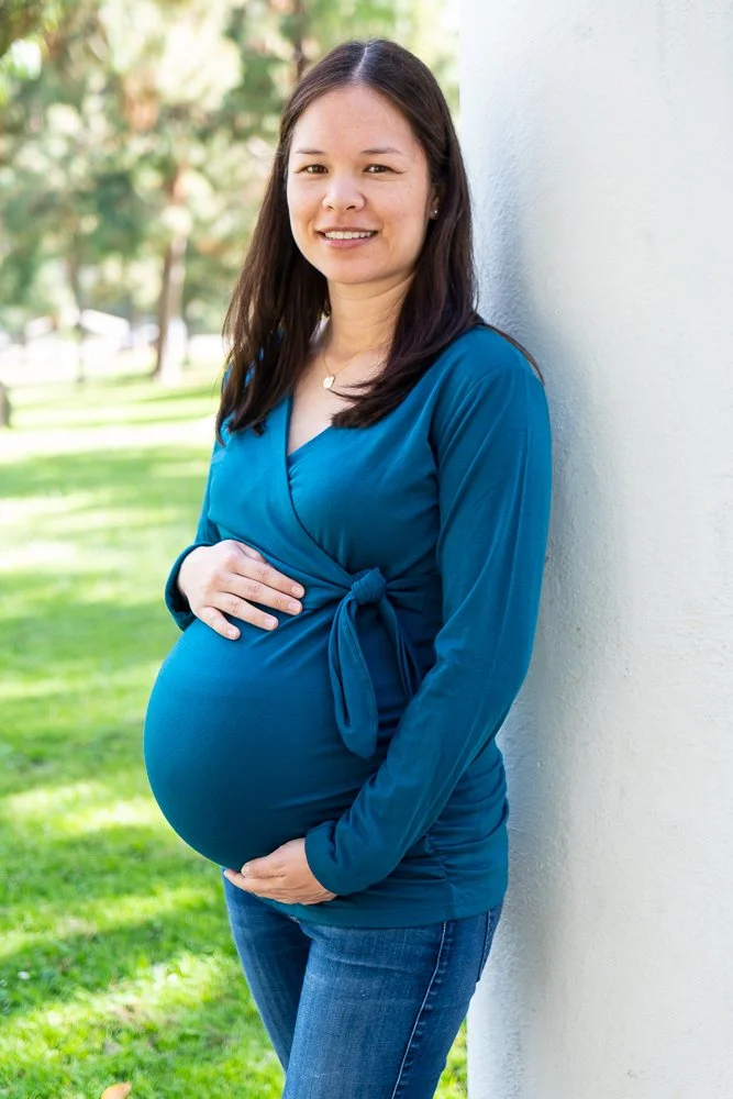 Woman with long dark hair wearing a blue dress, standing outdoors by a white wall, with her hand on her pregnant belly, smiling at the camera.