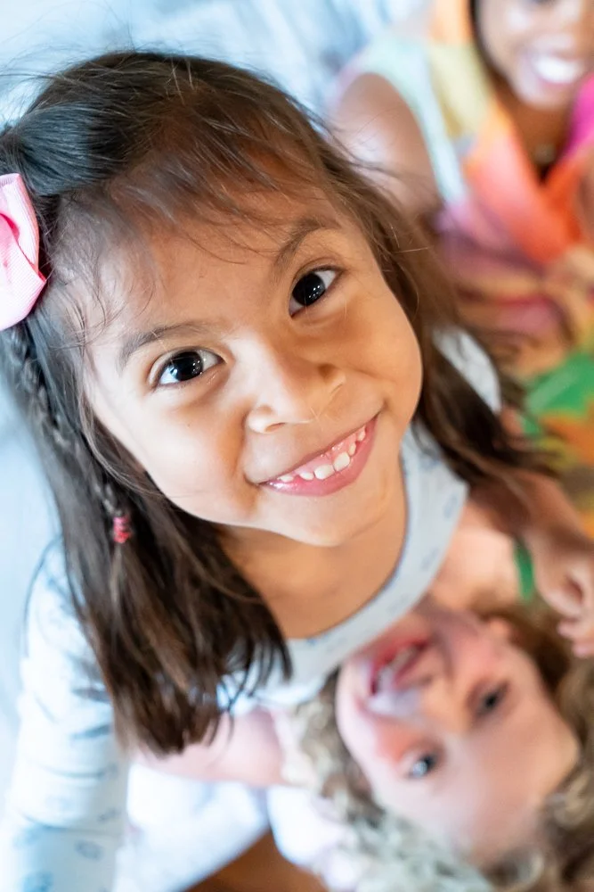 Close-up of two young girls smiling, one with a pink hair bow, lying on the floor, with a blurred background.