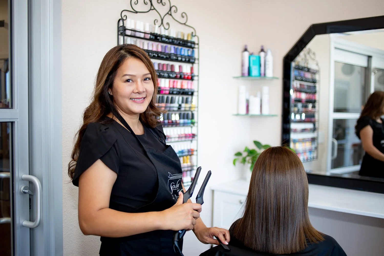 A smiling woman standing in a salon, holding hair styling tools, with a customer seated in front of a mirror. Shelves with hair products and a reflection of the woman in the mirror are also visible.