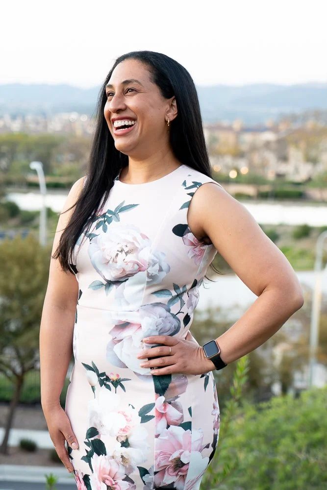 A woman with long black hair smiling outdoors in a sleeveless floral dress with pink and white flowers and green leaves, wearing a smartwatch on her left wrist, standing with her left hand on her hip.