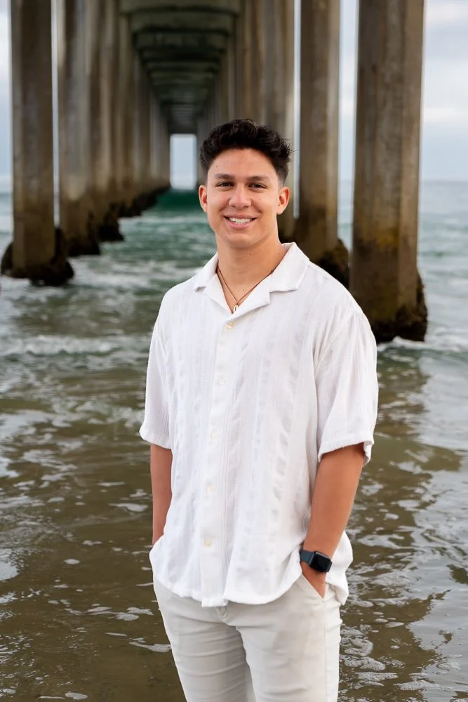 Young man standing in the ocean beneath a pier, smiling at the camera, wearing a white short-sleeve shirt and light-colored pants.