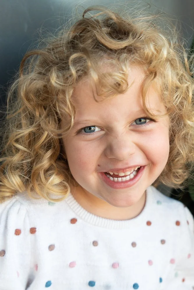 A smiling young girl with curly blonde hair and blue eyes, wearing a white sweater with multicolored polka dots.