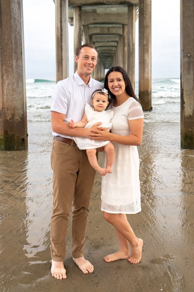 A family of three posing on a beach under a pier, with the ocean in the background. The man and woman are barefoot, and the woman is holding a young girl in a white dress.