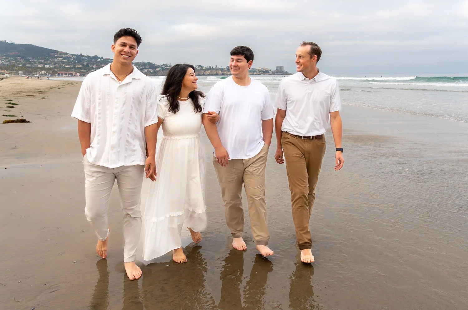 Group of four people walking on the beach, holding hands and smiling, dressed in white and khaki clothing, with ocean waves and a cityscape in the background.