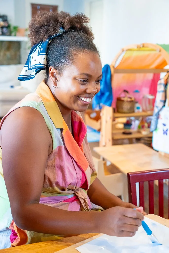 A woman with curly hair tied with a blue bandana, smiling while writing with a blue marker at a wooden table in a colorful, cozy indoor space.