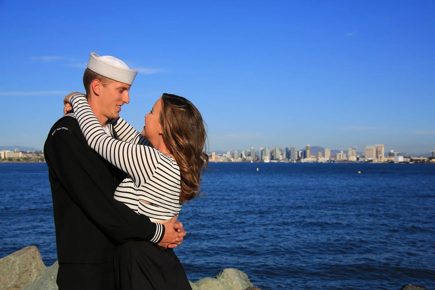 A couple embracing by the water with a city skyline in the background, under a clear blue sky.