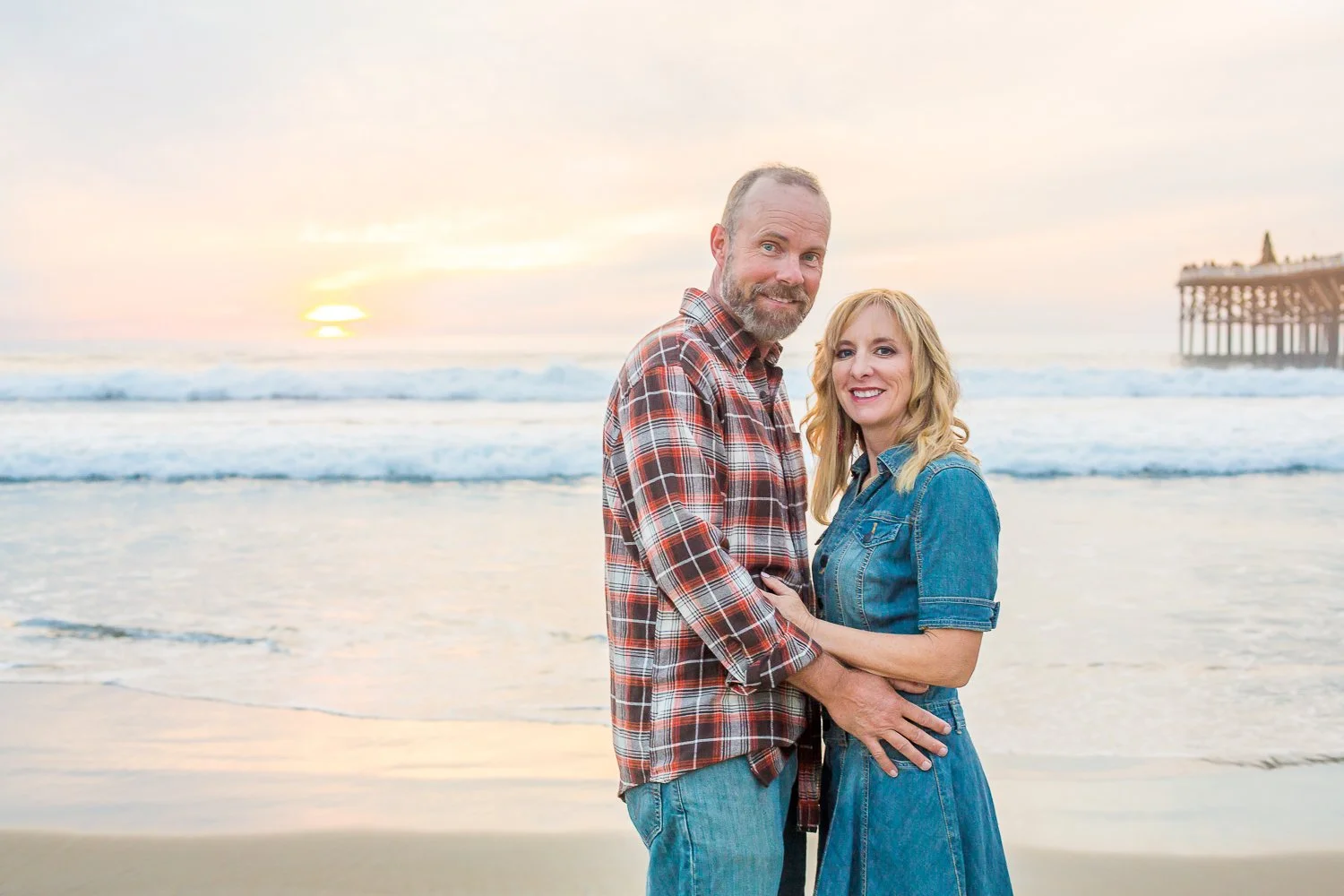A smiling couple stands on the beach at sunset, with ocean waves and a pier in the background.