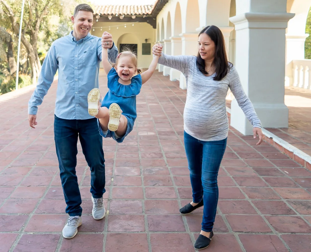 A family of three, a father, mother, and young daughter, enjoying an outdoor moment. The father is wearing a light blue shirt and jeans, the mother is wearing a gray striped shirt and jeans, and the daughter is in a blue dress. The daughter is being