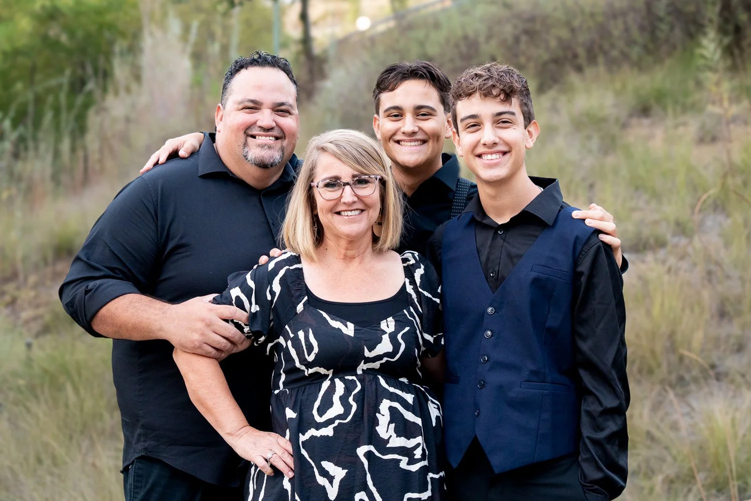 A family of five posing together outdoors. The background features grassy fields and trees. The family includes two adult men, one adult woman, and two teenage boys. They are all smiling and dressed in dark-colored semi-formal clothing.