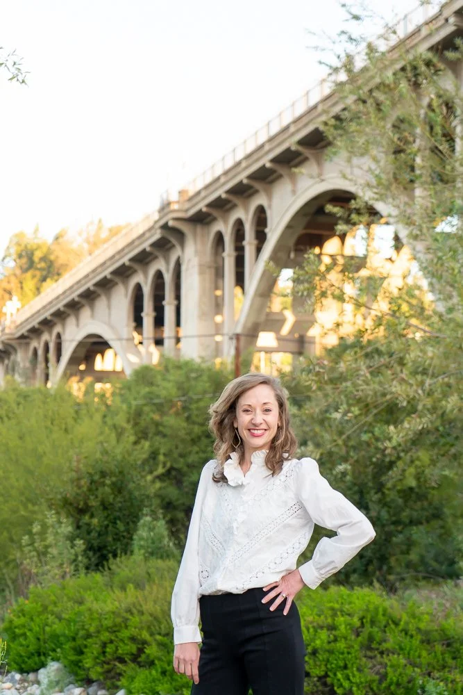 A woman wearing a white blouse and black pants standing outdoors with a bridge and greenery in the background.