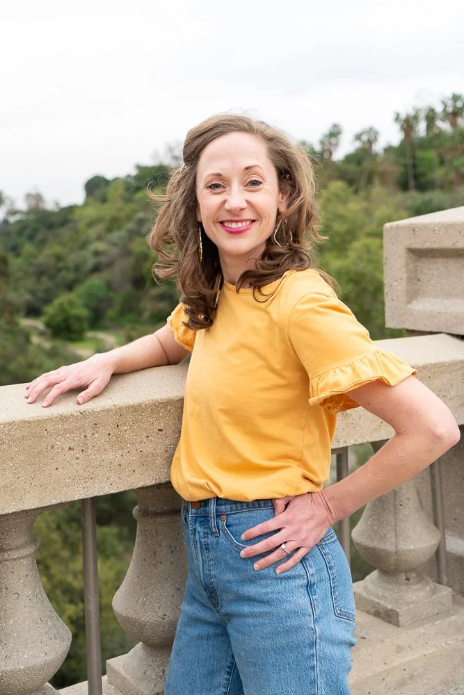 A woman with wavy brown hair smiling and wearing a yellow short-sleeve blouse and blue jeans, standing outdoors on a stone balcony.
